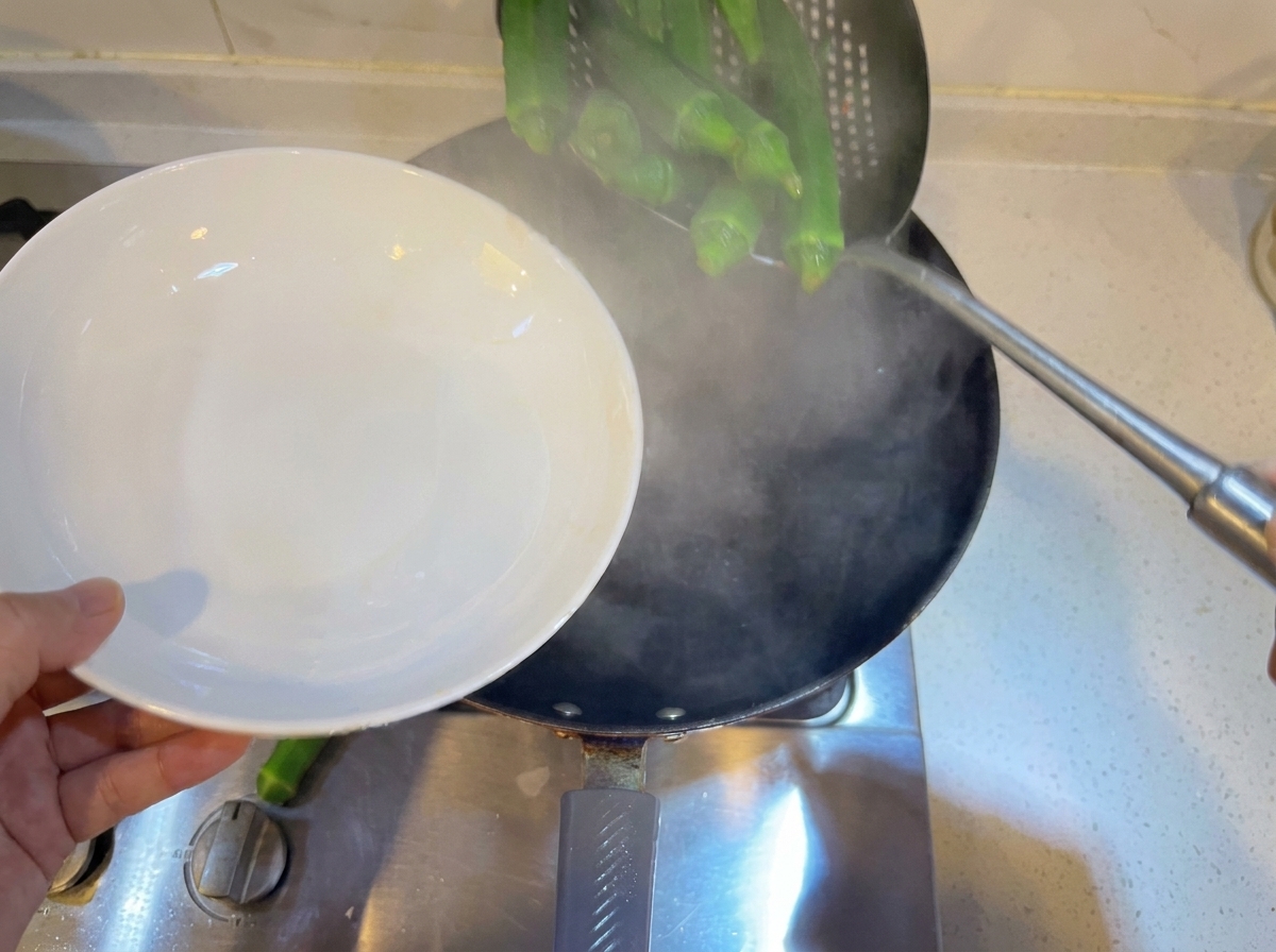 Okra verde intera sbollentata in una padella di acqua bollente usando un mestolo forato.