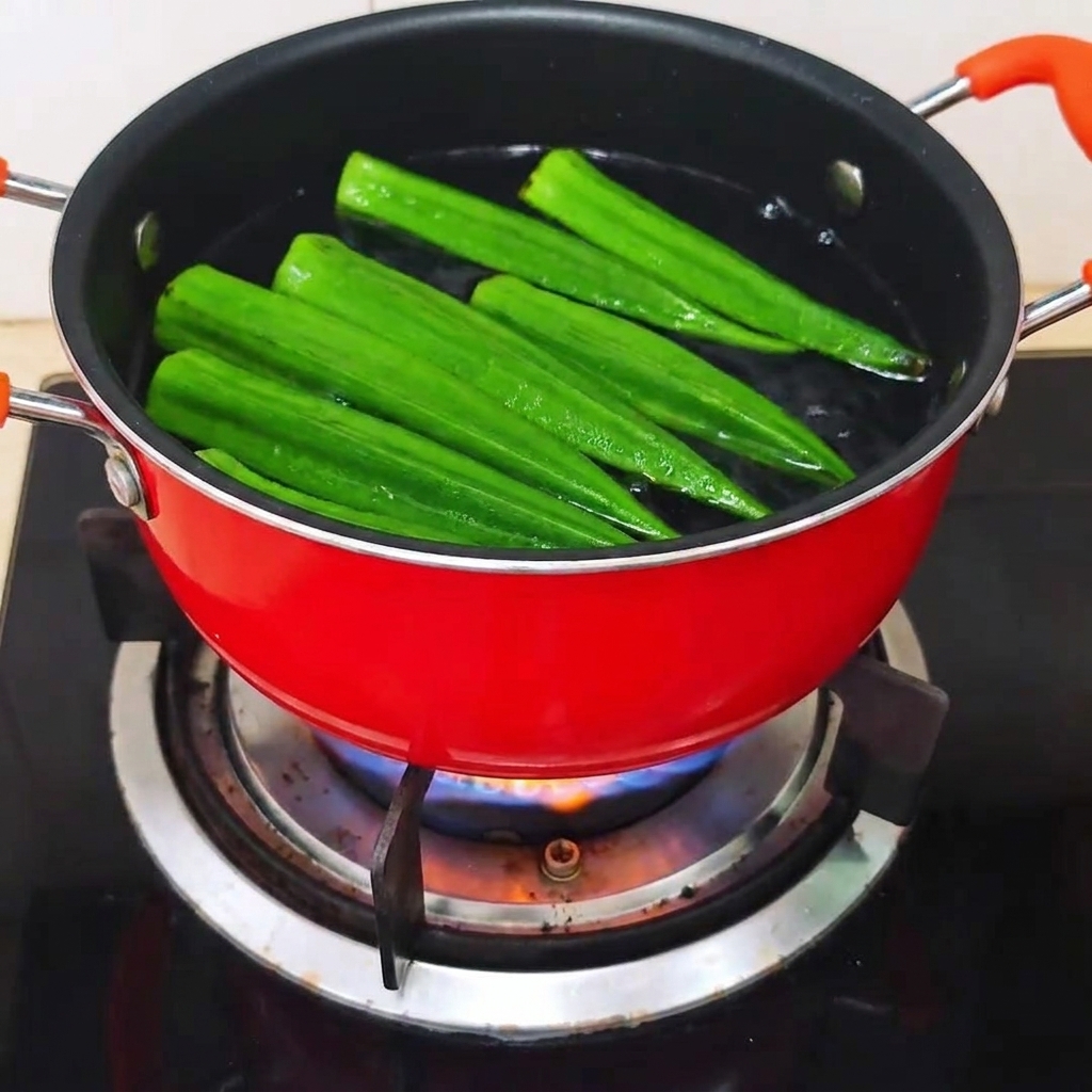 Interi baccelli di okra cruda inseriti in una pentola rossa di acqua bollente.