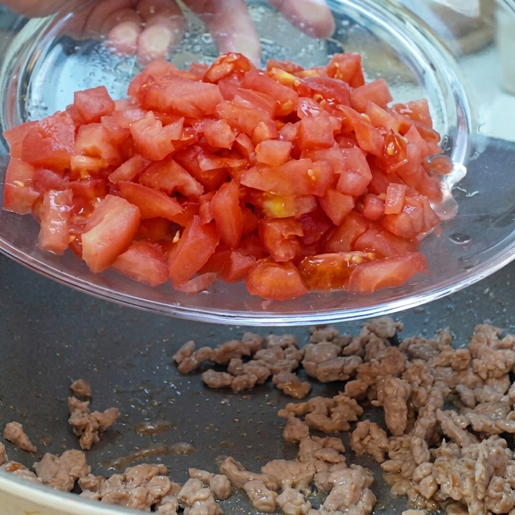 Pomodori a dadini e cipolle rosse versati da una ciotola di vetro nella padella con la carne.