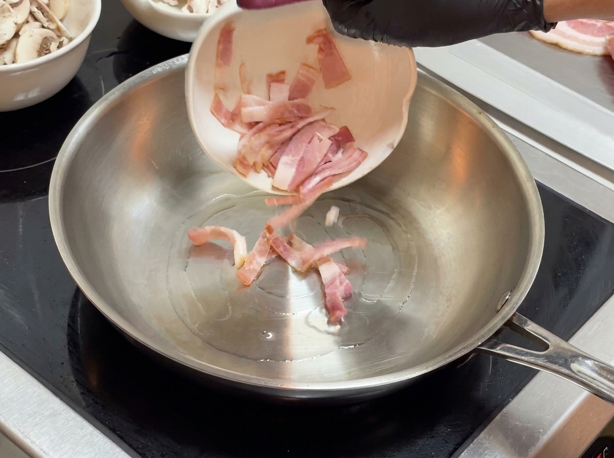 Pezzi di pancetta versati da una ciotola bianca piccola in una padella di acciaio inossidabile.
