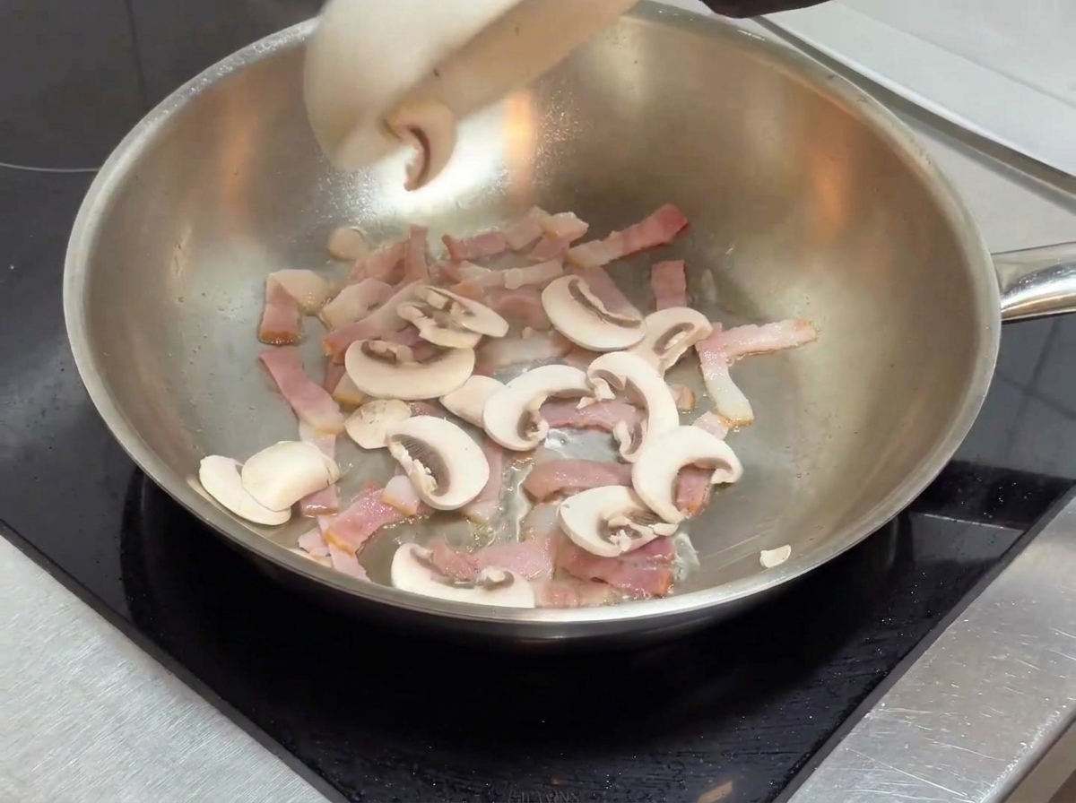 Funghi affettati aggiunti alla padella contenente pezzi di pancetta cotta.