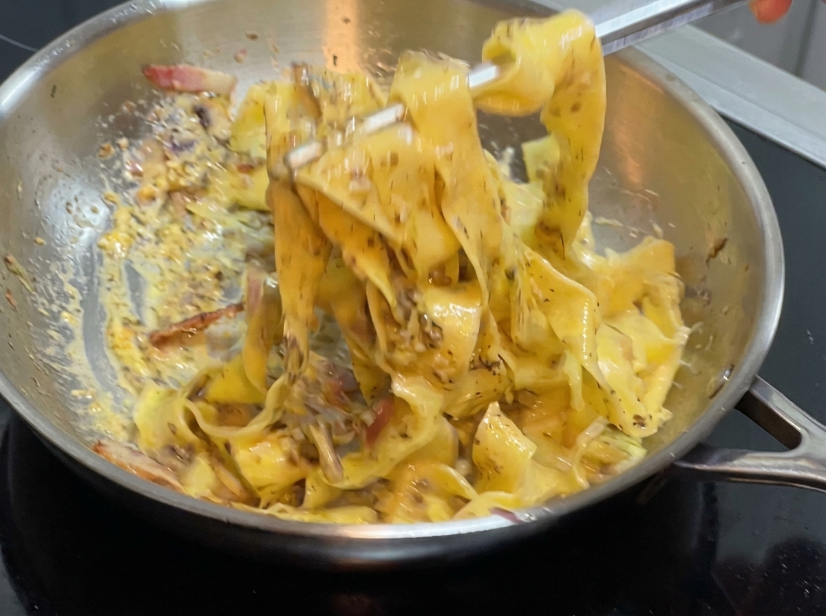 Un paio di pinze che mescolano pasta larga e salsa allinterno di una padella in acciaio inossidabile sul fuoco.