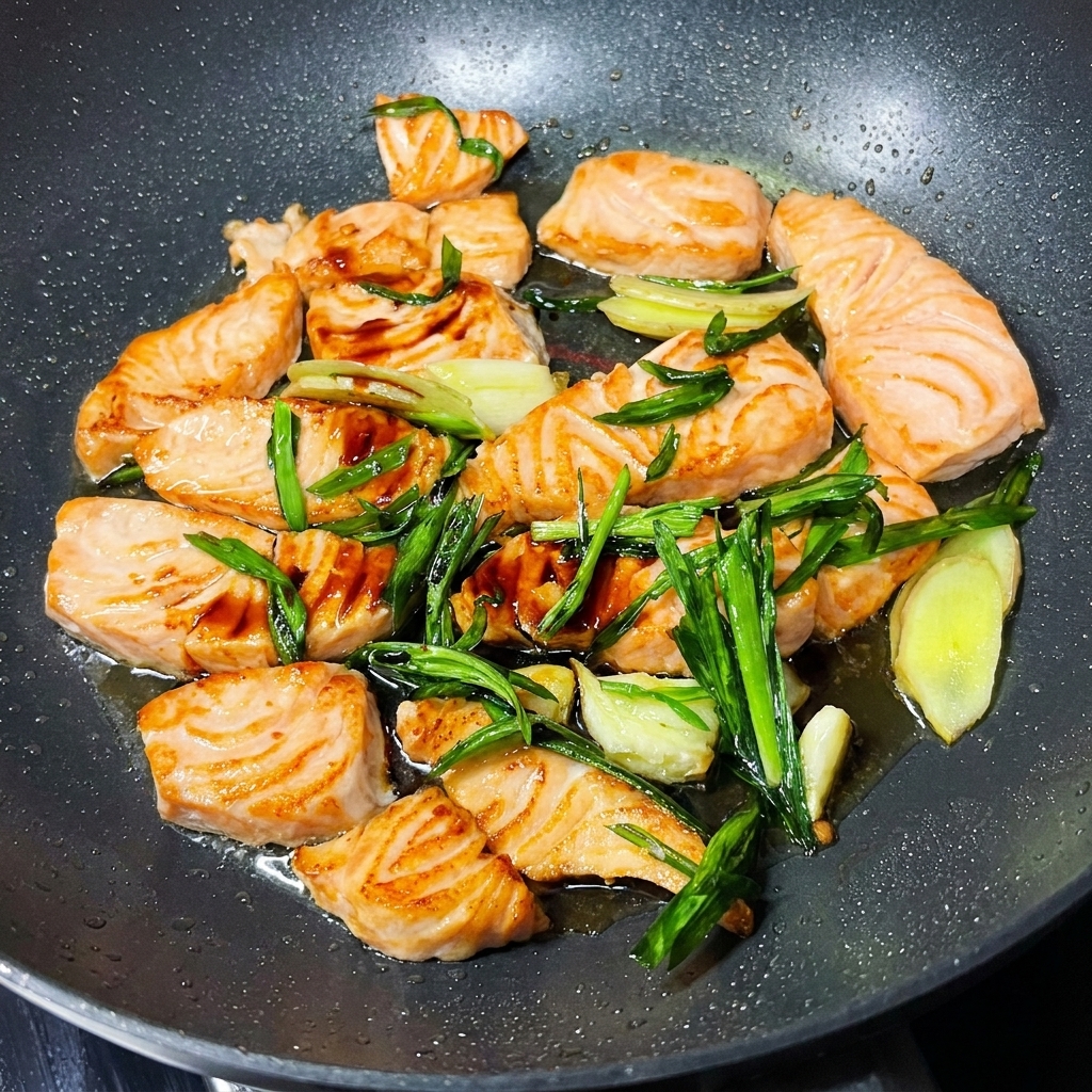 Pezzi di salmone in padella ricoperti di salsa di soia e guarniti con cipollotti verdi freschi e semi di sesamo.