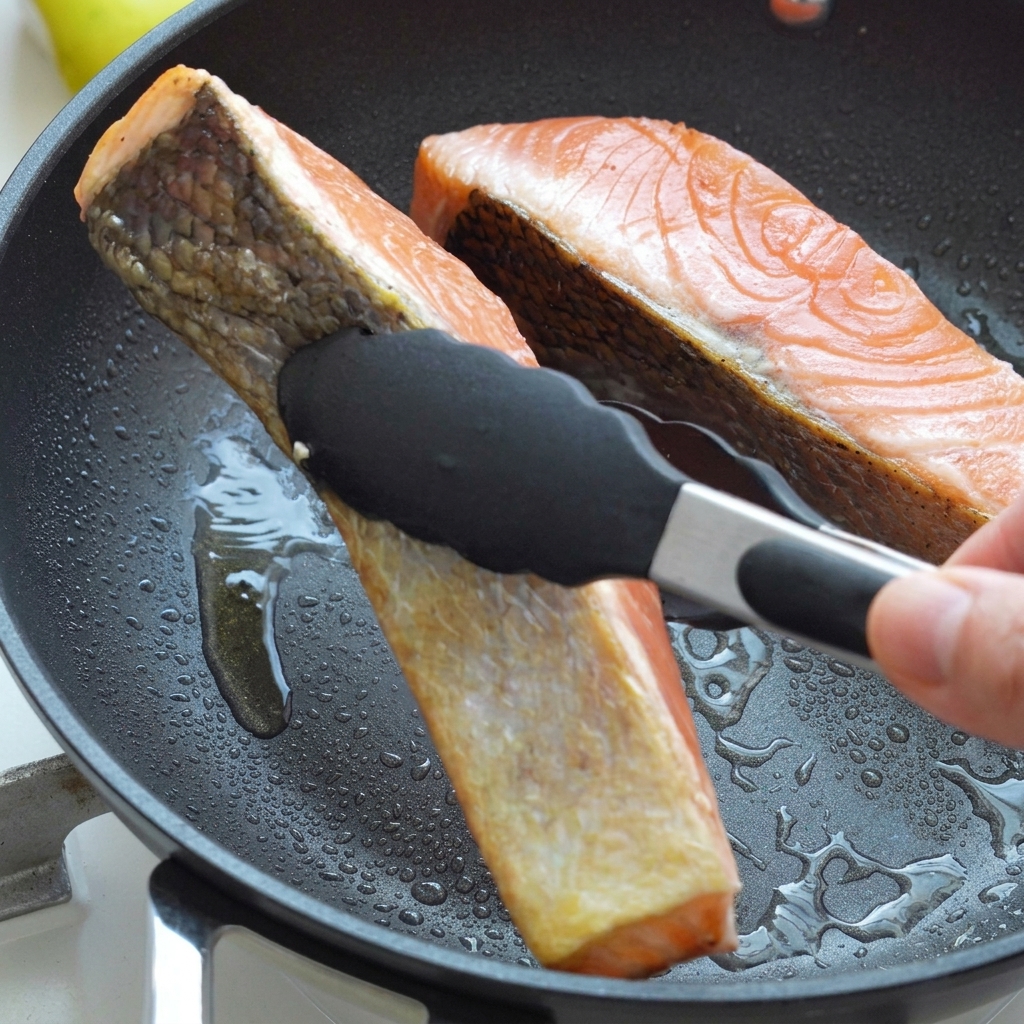 Filetti di salmone girati con pinze nere in una padella calda, che mostrano una pelle ben dorata e croccante.