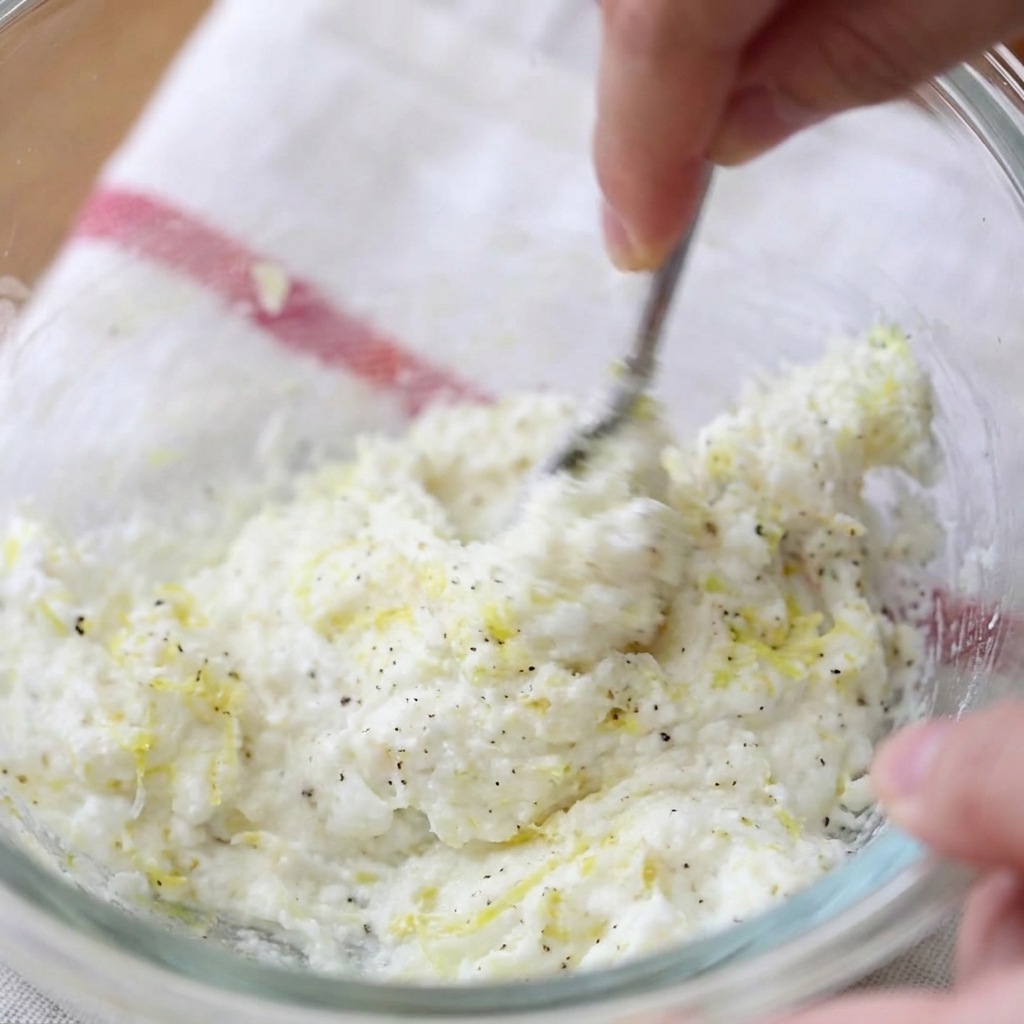 Una mano che usa una فورك (forchetta) per mescolare formaggio spalmabile, scorza di limone e pepe nero in una ciotola di vetro.