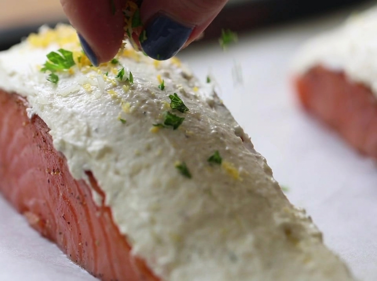 Dita che cospargono una miscela di pangrattato ed erbe verdi e gialle sopra filetti di salmone crudo ricoperti di formaggio spalmabile bianco.