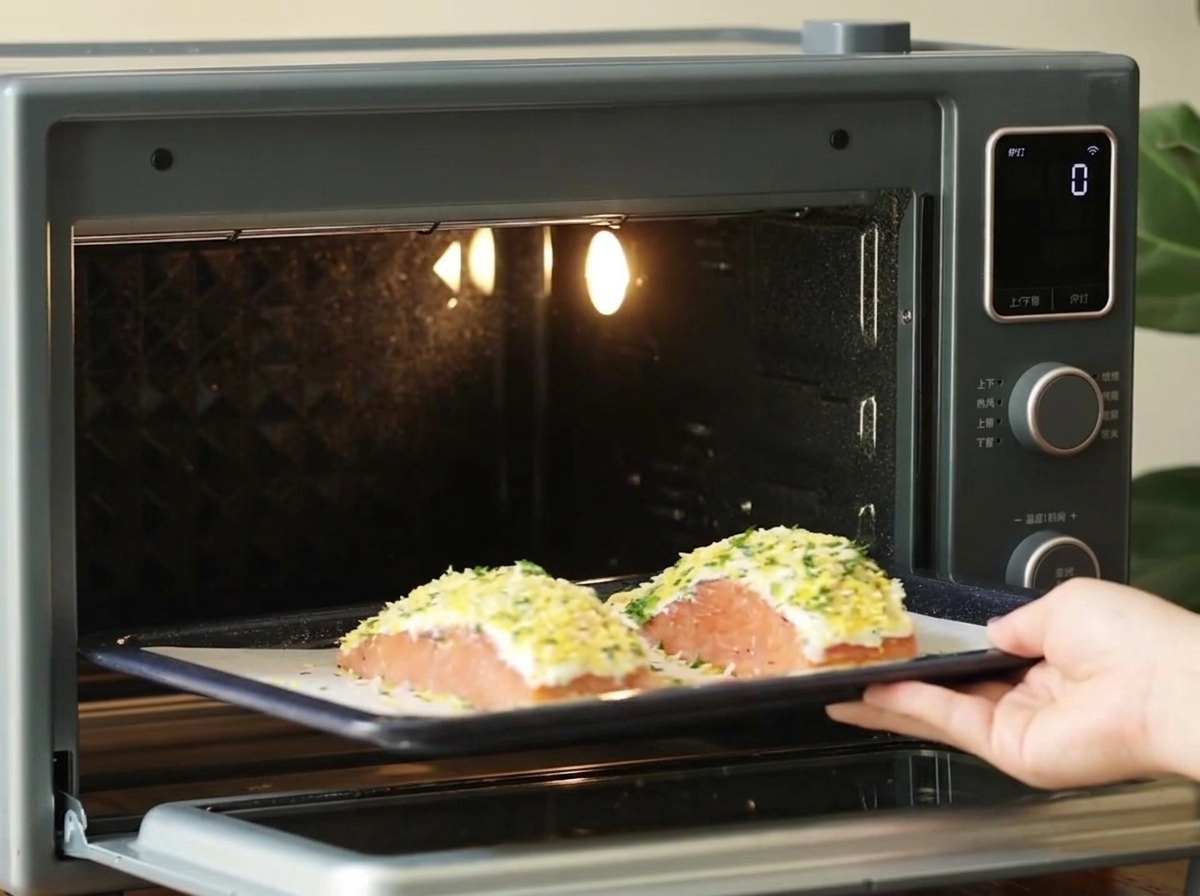 Una mano che fa scorrere una teglia scura con due filetti di salmone impanati in un piccolo forno da banco fortemente illuminato.