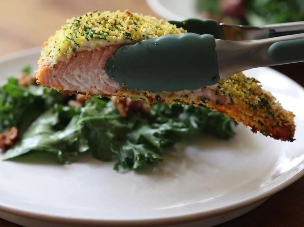 Pinze da cucina in silicone verde che reggono un pezzo di salmone al forno con una crosta dorata e croccante sopra un piatto bianco contenente uninsalata di cavolo verde.