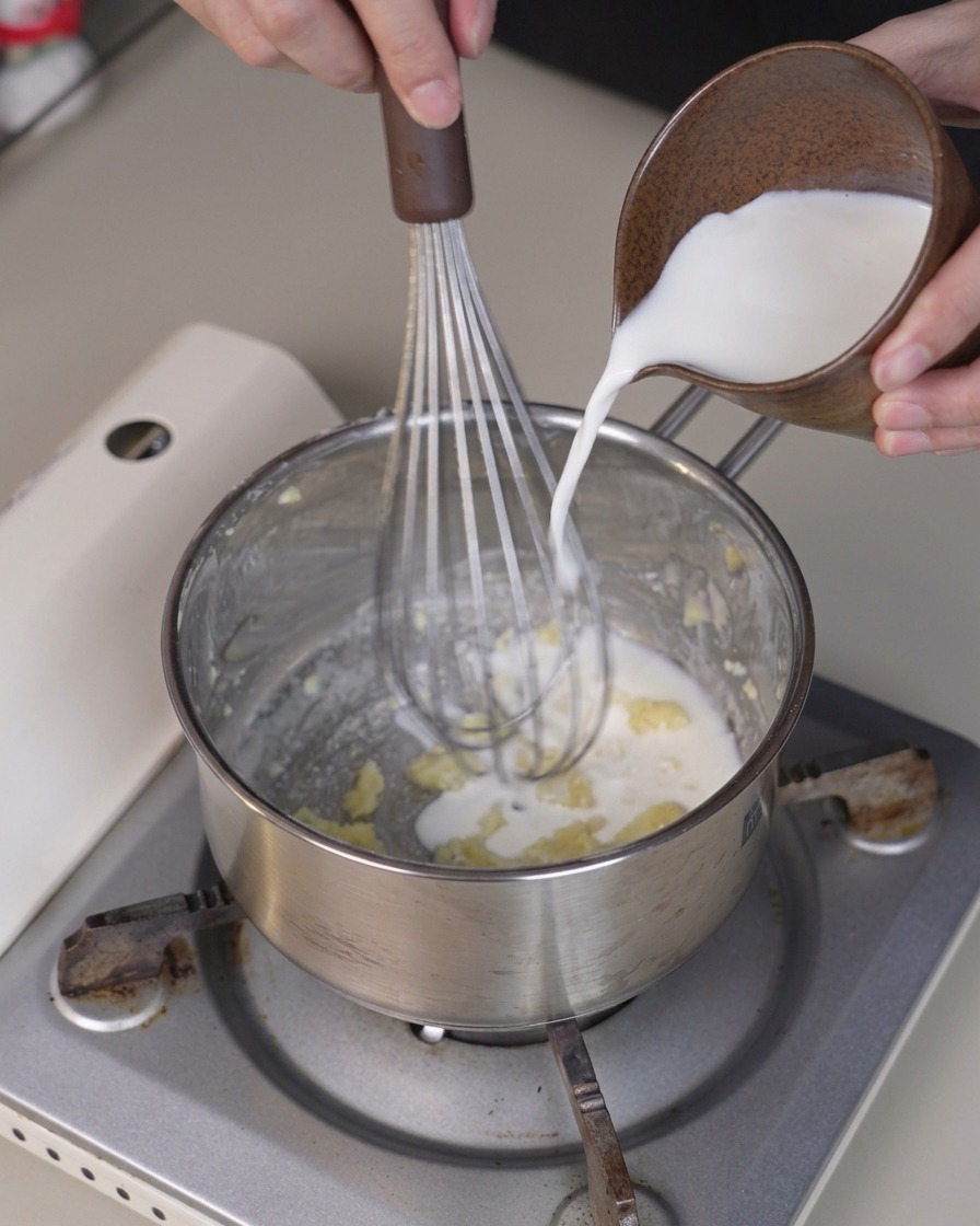 Latte freddo versato da una piccola brocca in un pentolino mentre una frusta mescola il composto.