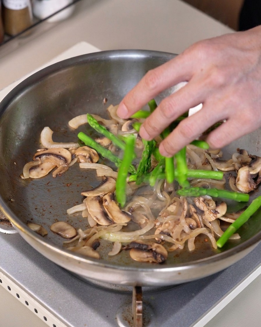 Pezzi di asparagi verdi mescolati nella padella contenente cipolle e funghi saltati.