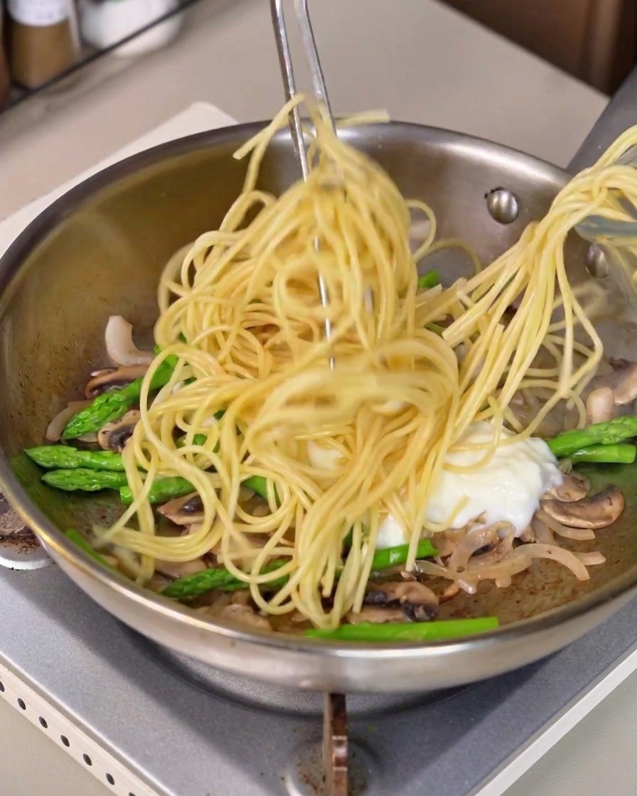 Spaghetti lunghi sollevati con le pinze e mescolati nella padella con salsa bianca e verdure.