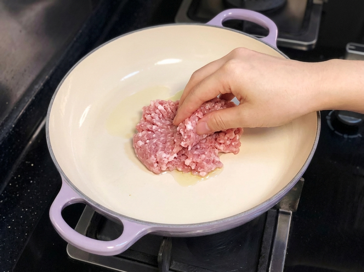 Una mano mette la carne di maiale macinata in una padella con manico viola contenente olio doliva.