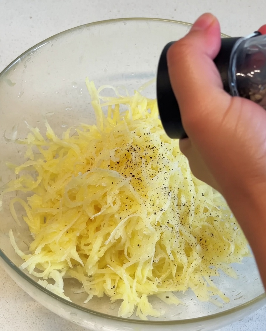 Patate gialle grattugiate allinterno di una ciotola di vetro che vengono condite con pepe nero da un macinino manuale.