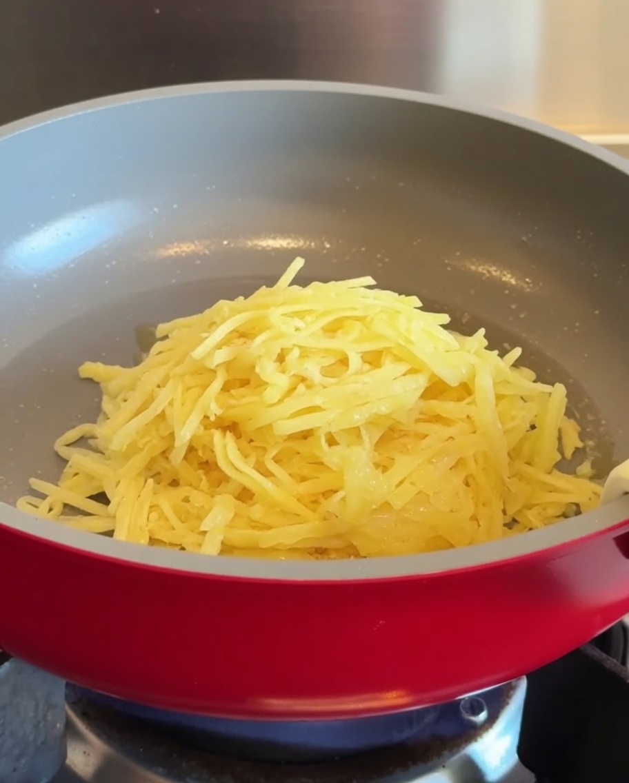 Un mucchietto di patate grattugiate posizionato al centro di una padella antiaderente rossa ben calda.