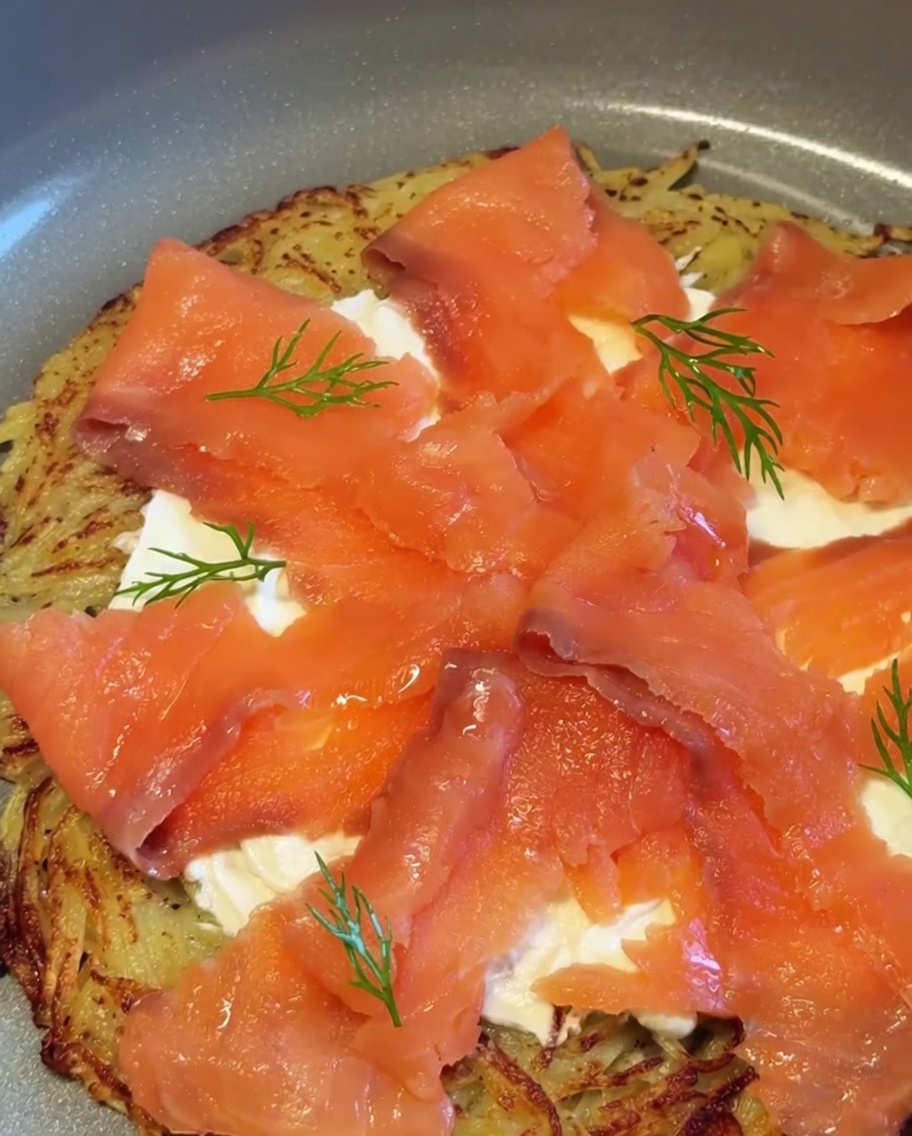 Una pizza di patate e salmone affumicato finita, guarnita con rametti di aneto fresco e pezzetti di cipolla rossa.