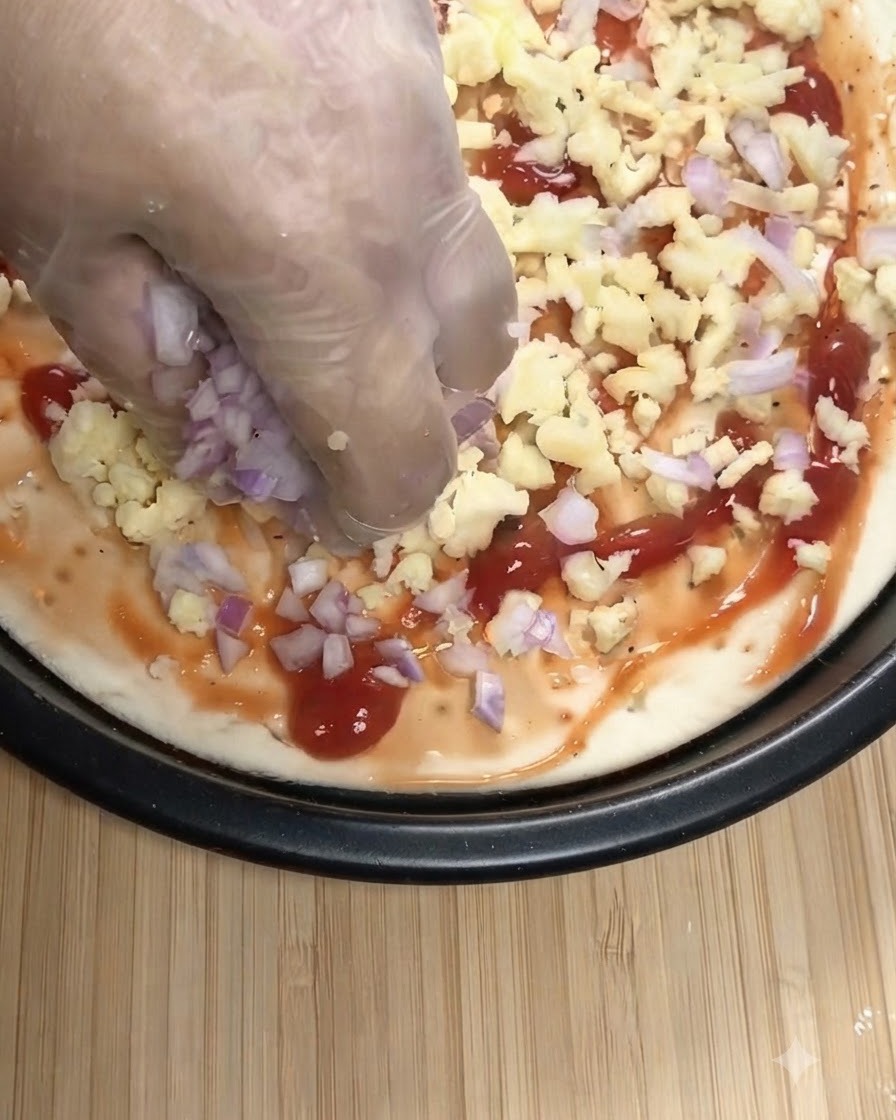 Una mano con un guanto trasparente che sparge cipolle rosse tritate e mozzarella grattugiata su una base per pizza coperta di salsa di pomodoro.