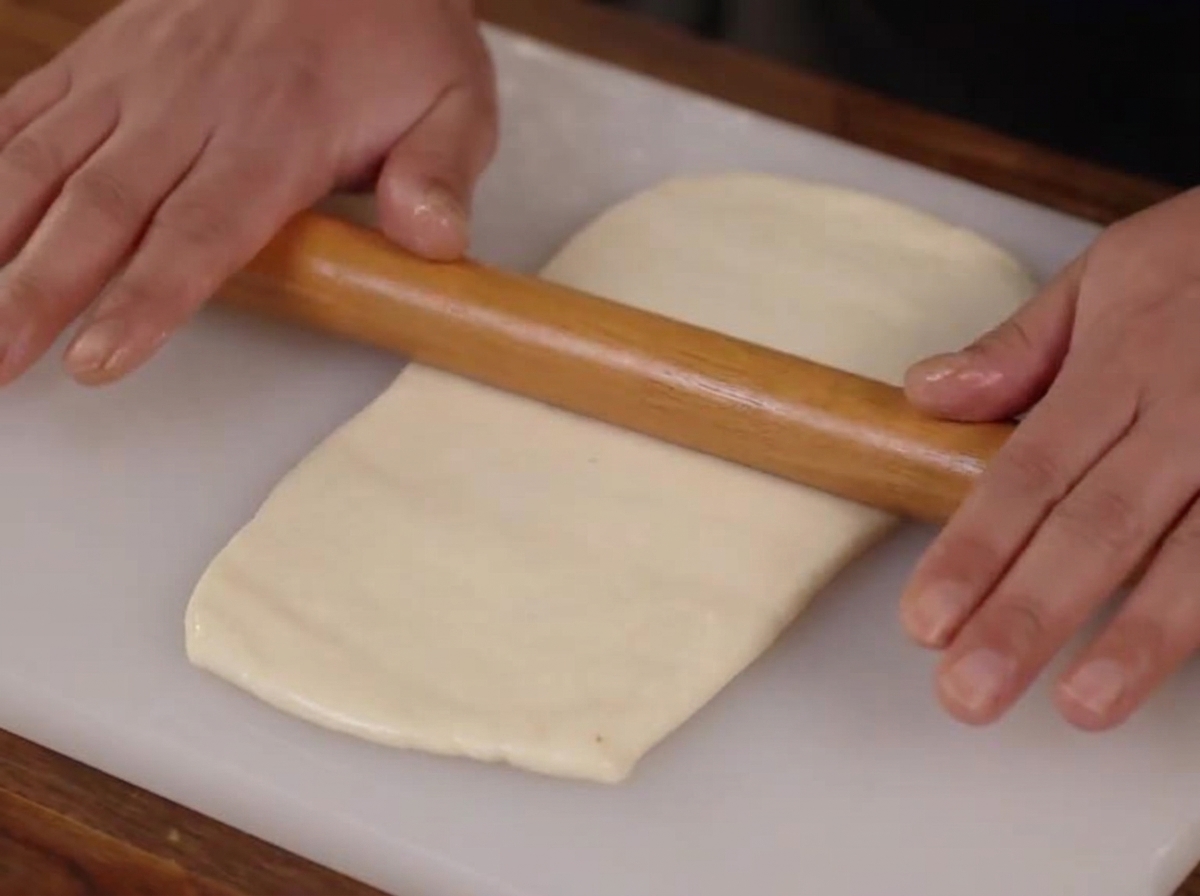 Mani che usano un mattarello di legno per appiattire un pezzo di impasto liscio in un rettangolo su un tagliere bianco.