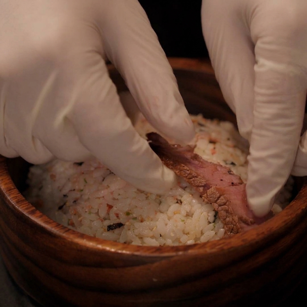 Mani con i guanti che posizionano fette di bistecca cotta sopra una ciotola di riso condito.
