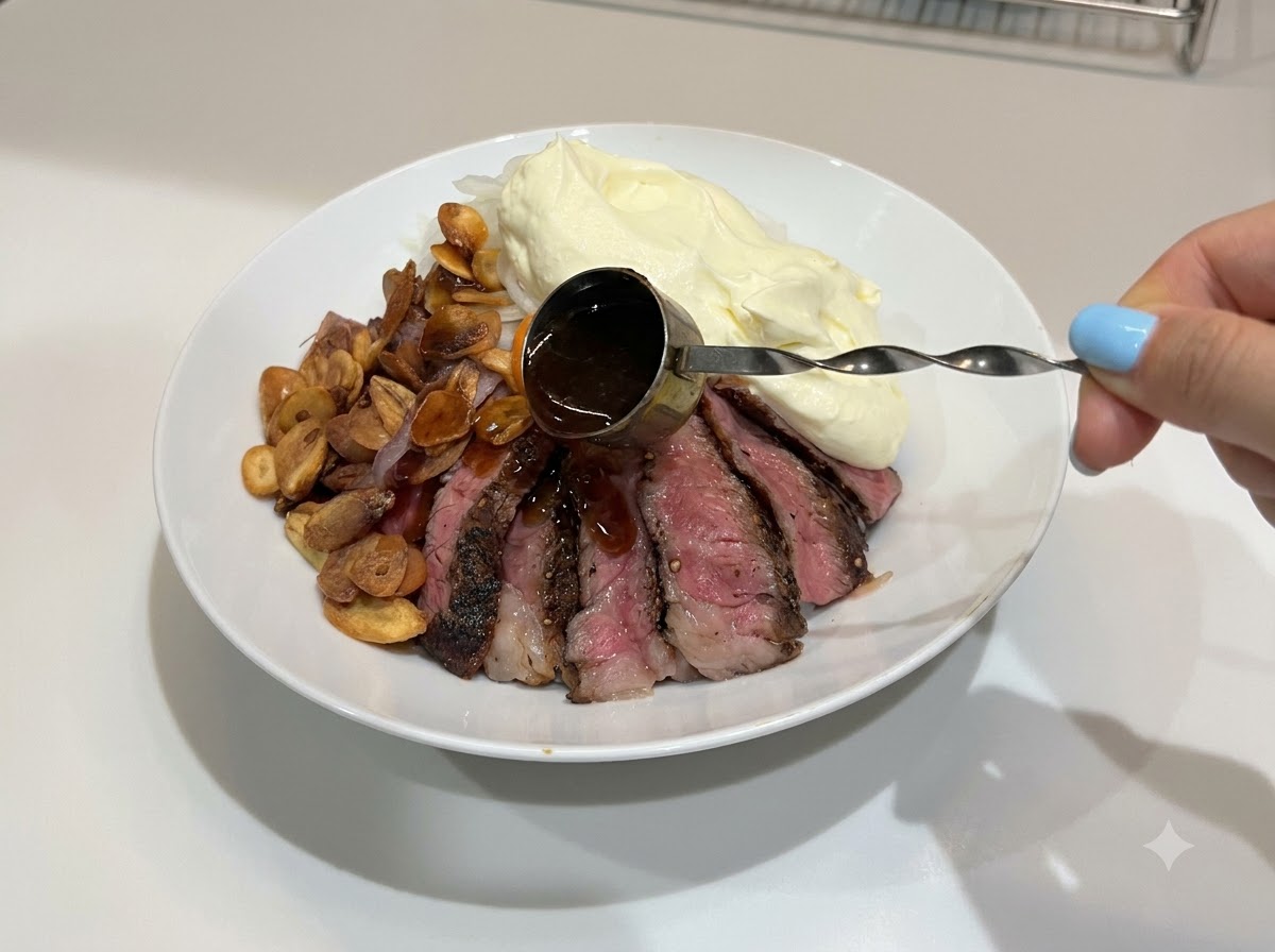 Una mano che utilizza un piccolo misurino con manico per irrorare salsa teriyaki scura e lucida sopra una ciotola di riso e bistecca completata.