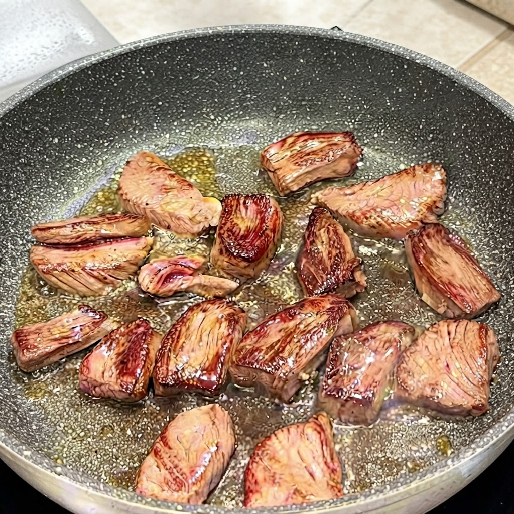 Pinze che girano i pezzi di bistecca di scamone in padella mostrando le superfici dorate.
