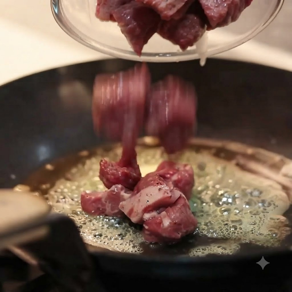 Pezzi di manzo crudo condito versati da una ciotola di vetro in una padella calda a fondo piatto piena di burro fuso e sfrigolante.