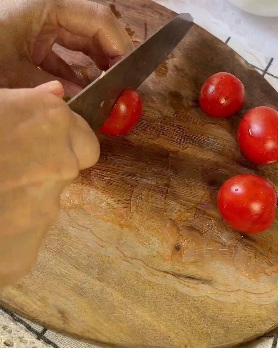 Un coltello che taglia un piccolo pomodoro rosso su un tagliere di legno accanto ad altri pomodori dimezzati.