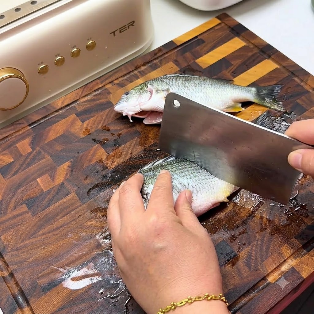 Una mano che usa una grande mannaia di metallo per praticare tagli diagonali sul lato di un pesce fresco su un tagliere di legno.