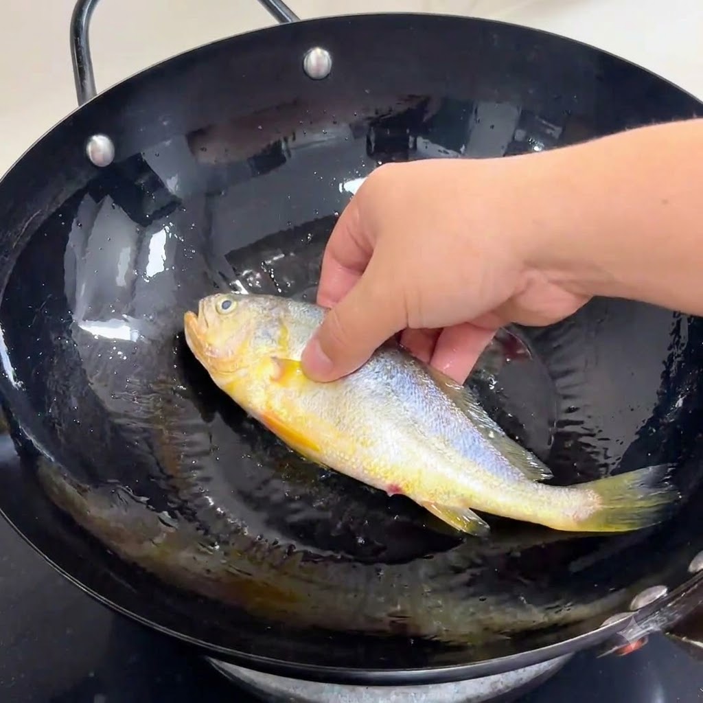 Una mano che adagia delicatamente un pesce intero condito in un wok nero pieno di olio ben caldo.