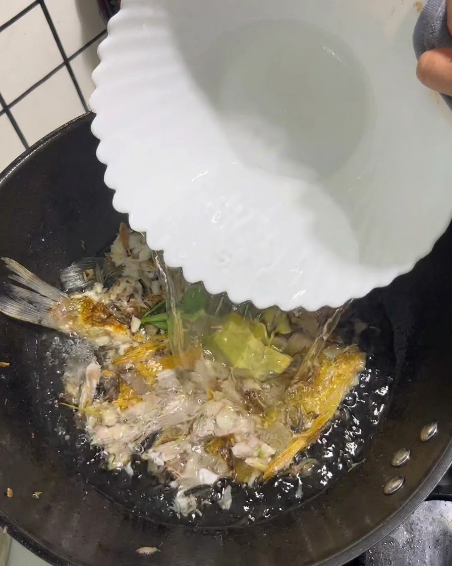 Acqua bollente limpida versata da una ciotola bianca in un wok contenente pesce schiacciato e aromi.
