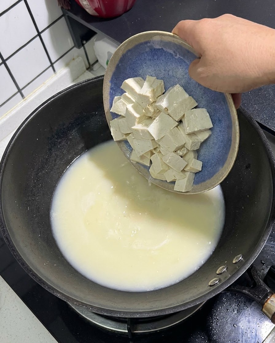Cubetti di tofu bianco che scivolano da un piatto blu in una pentola di brodo di pesce bianco e cremoso.
