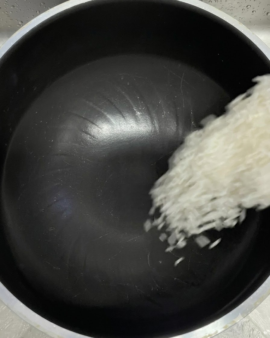 Chicchi di riso bianco che cadono da un misurino in una pentola interna nera di un cuociriso.