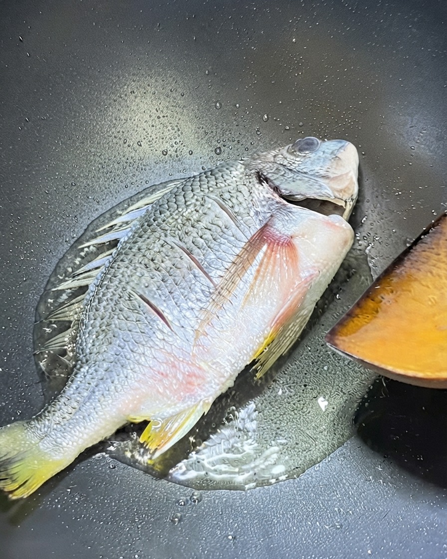 Un intero dentice giallo che cuoce in un wok con una spatola di legno posizionata vicino per girarlo.