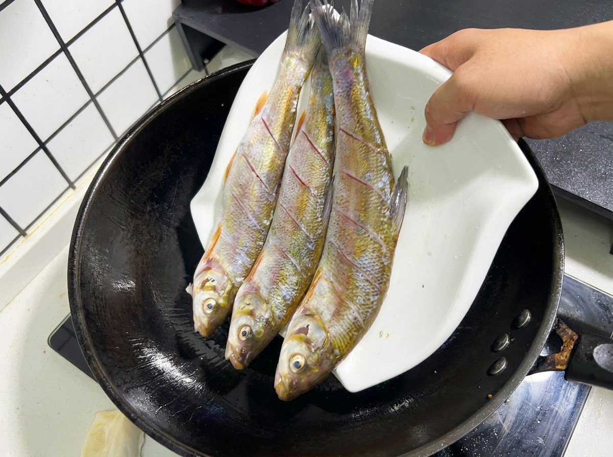 Tre pesci interi, incisi e crudi che vengono fatti scivolare da un piatto bianco in un wok nero con olio caldo.