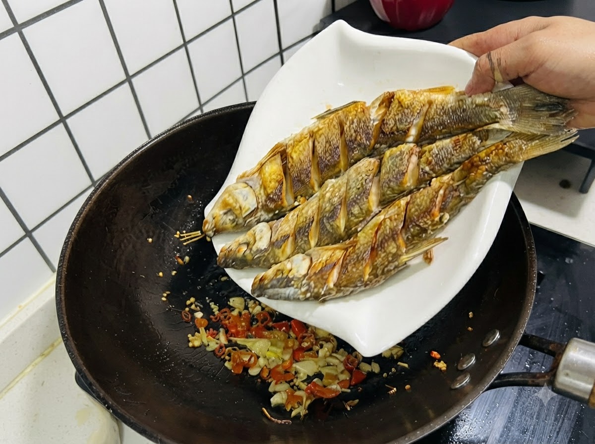 Tre pesci fritti fatti scivolare da un piatto bianco in un wok contenente aglio, zenzero e peperoncini saltati.