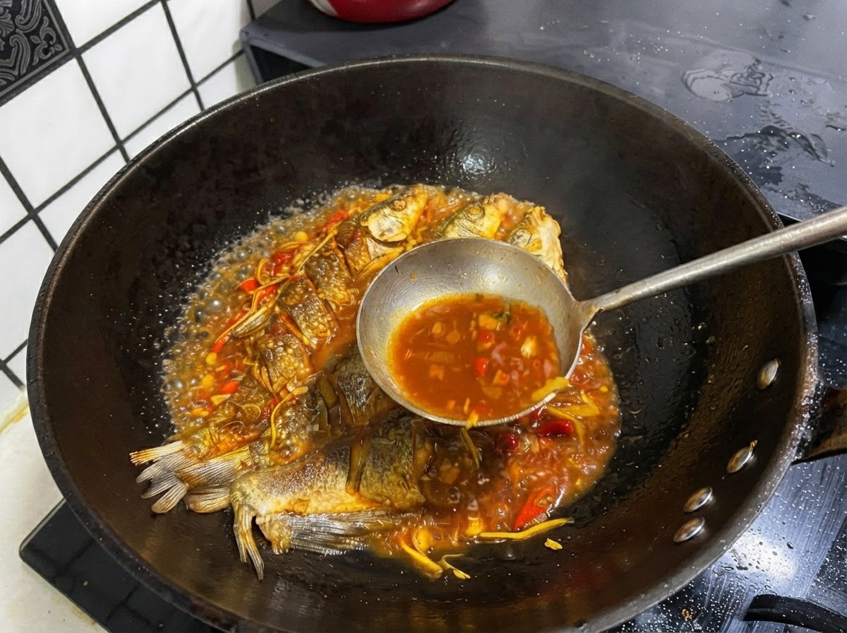 Un mestolo di metallo che raccoglie brodo scuro e saporito da una padella contenente pesce intero fritto.