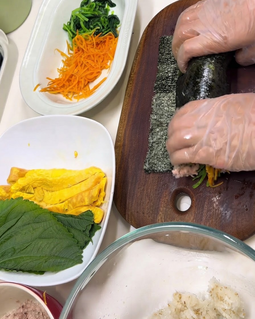 Mani che indossano guanti trasparenti avvolgono gli ingredienti allinterno di un foglio di alga nori su un tagliere di legno.