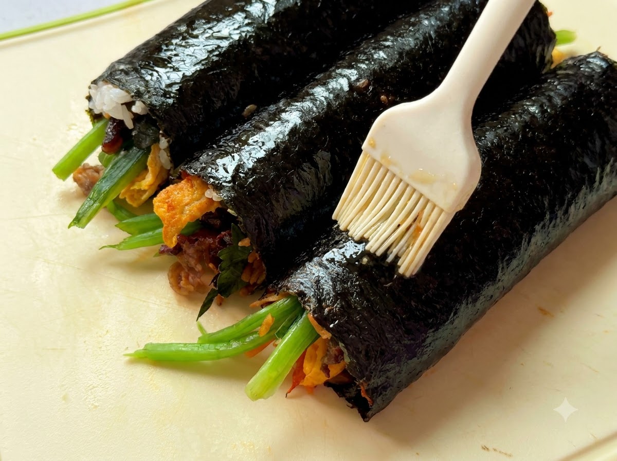 Primo piano dellolio di sesamo spennellato su tre rotoli di gimbap posizionati su un tagliere.