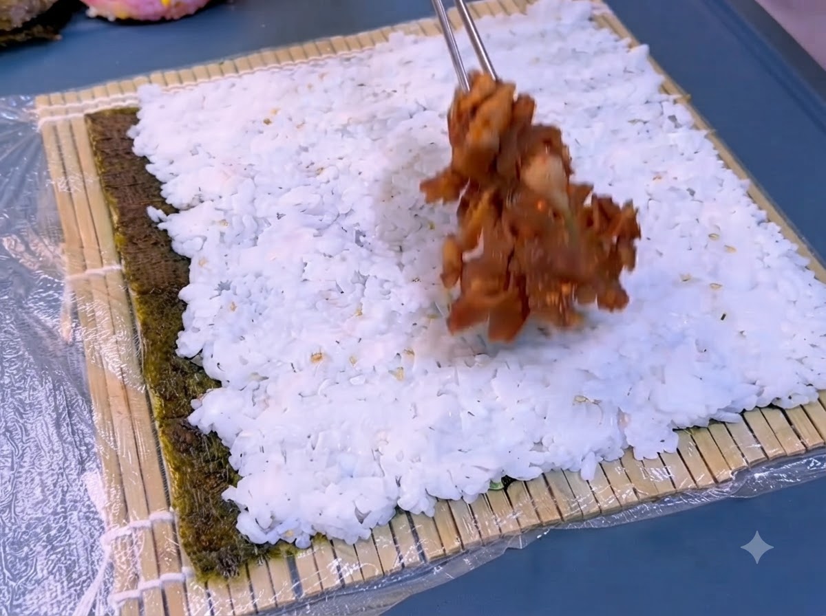 Un foglio di alga scura posizionato su un tappetino di bambù con uno strato di riso bianco cotto che viene distribuito sopra.