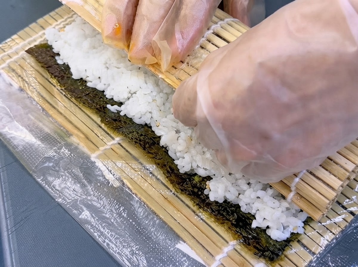 Mani con guanti che sollevano un tappetino di bambù per arrotolare strettamente alga e ripieno in un cilindro.