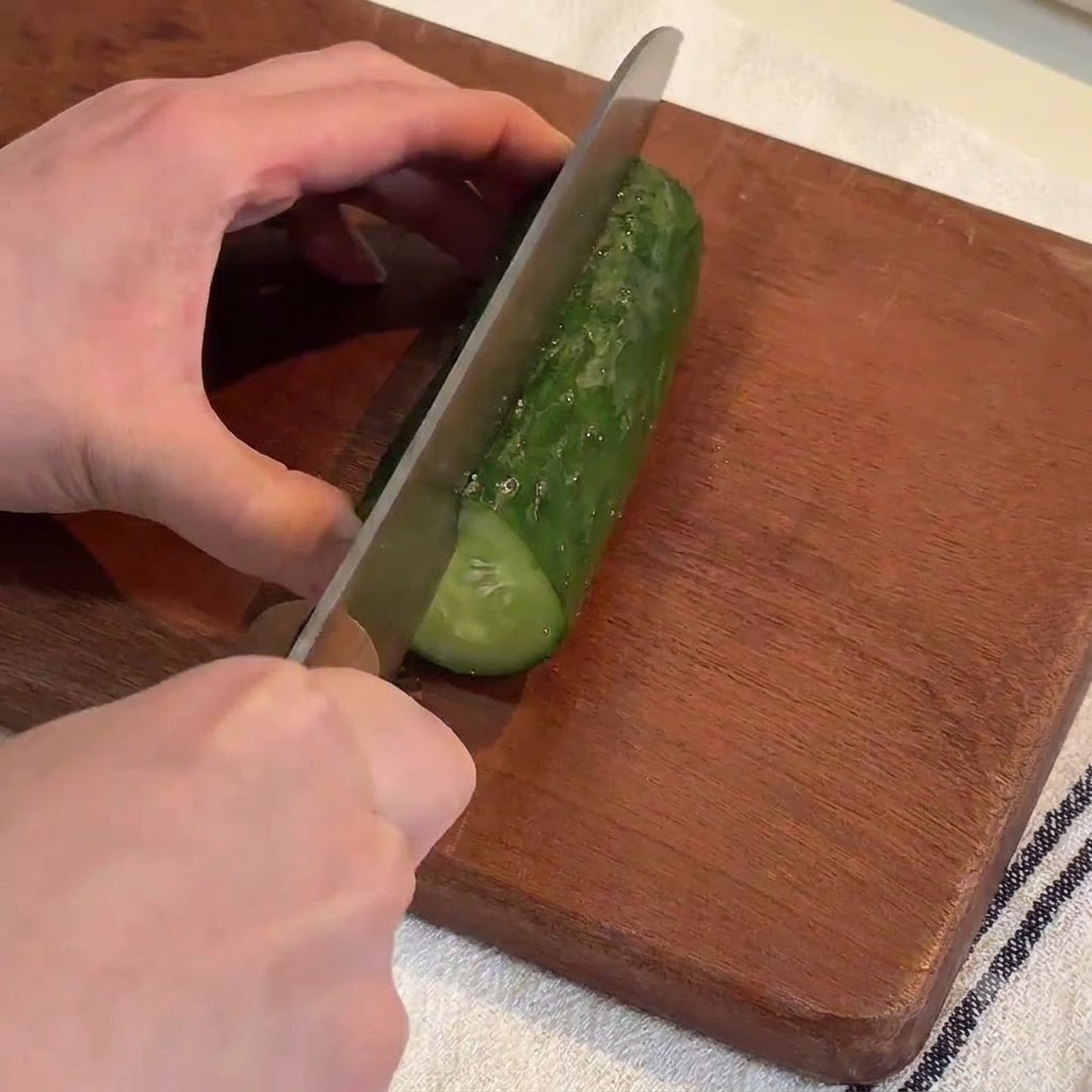 Mani che usano un coltello grande per affettare un cetriolo verde fresco in strisce lunghe su un tagliere di legno.