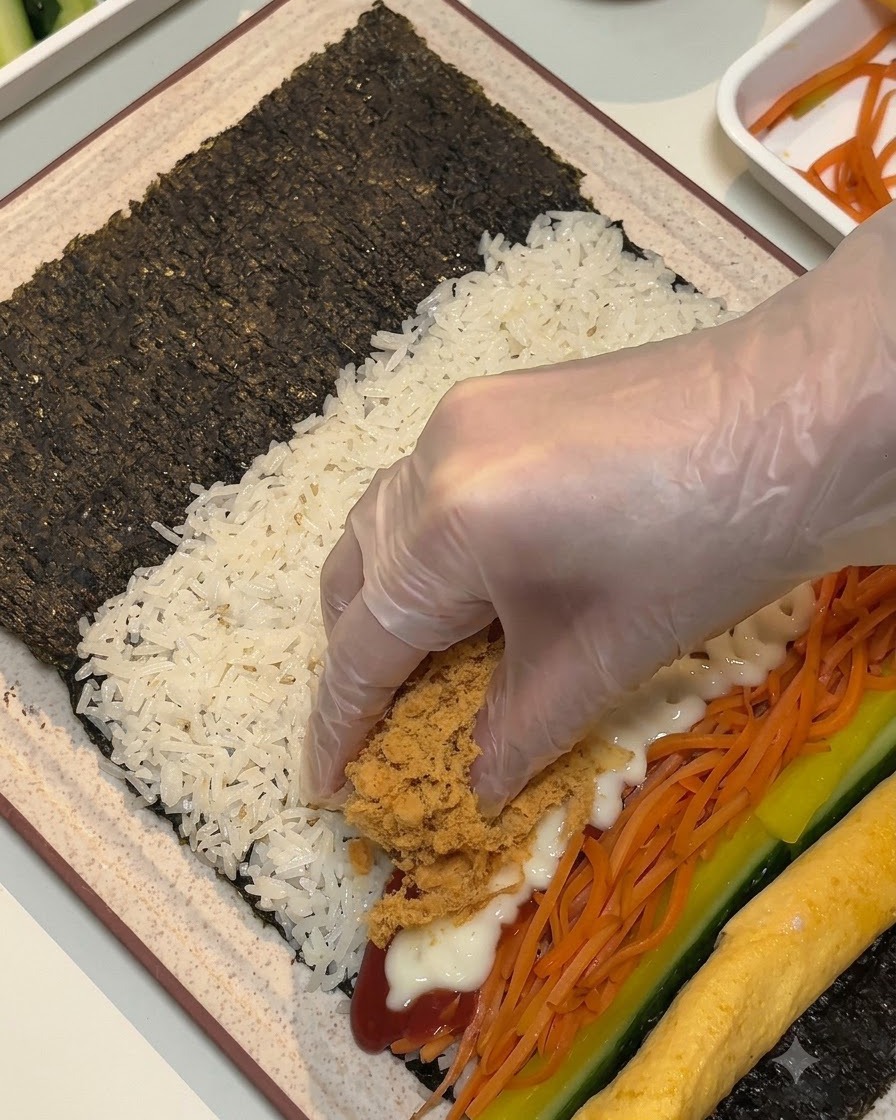 Una mano guantata che posiziona maiale sfilacciato marrone soffice e strisce di carne in scatola sui ripieni del foglio di alga.