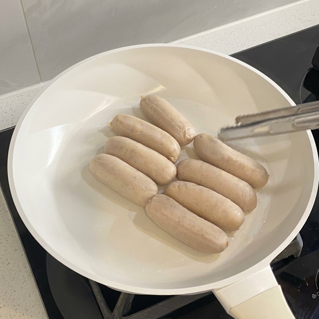 Salsicce che cuociono e dorano uniformemente in una padella bianca.