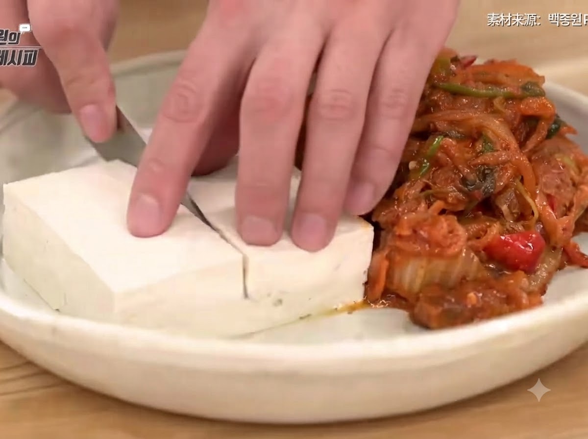 Una mano che usa un coltello per affettare ordinatamente il tofu sbollentato su un piatto accanto a un mucchietto di saporito kimchi saltato e maiale.