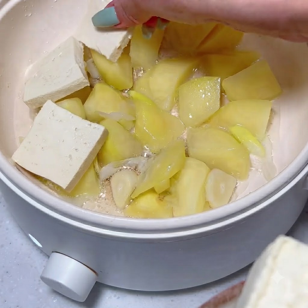 Fette di tofu disposte ordinatamente sopra uno strato di pezzi di patata cotti e aglio dentro una pentola.