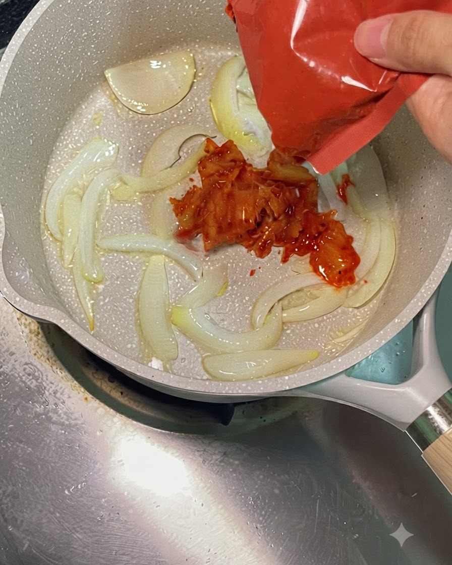 Kimchi tritato rosso vivo aggiunto da una confezione rossa in una padella calda con cipolle affettate saltate.