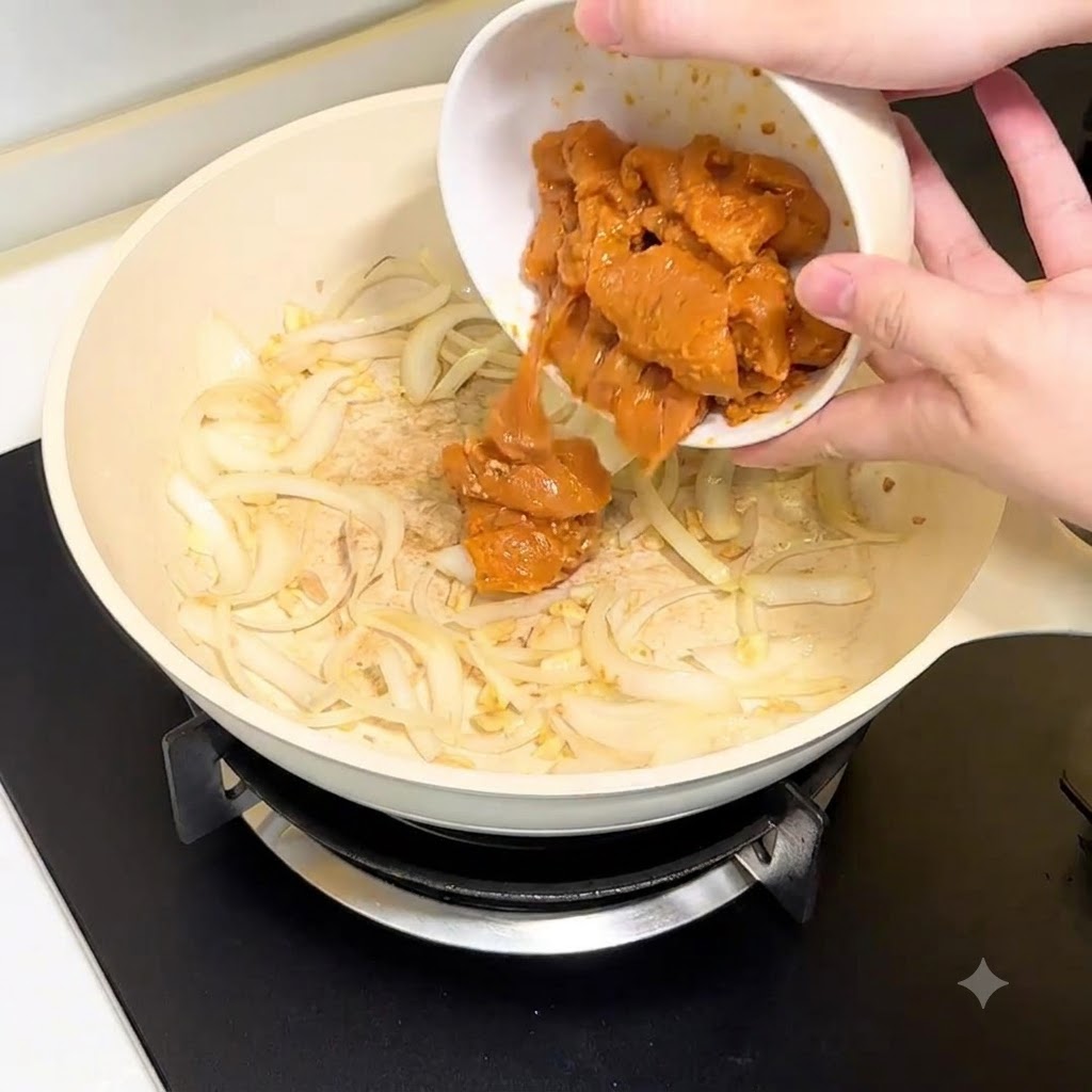 Pezzi di petto di pollo versati da una ciotola bianca nella padella con cipolle e aglio.