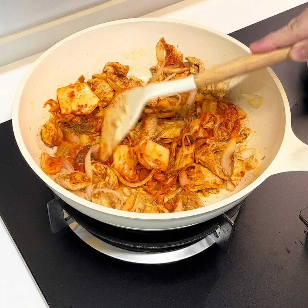 Kimchi e pollo saltati insieme in una padella bianca con una spatola.