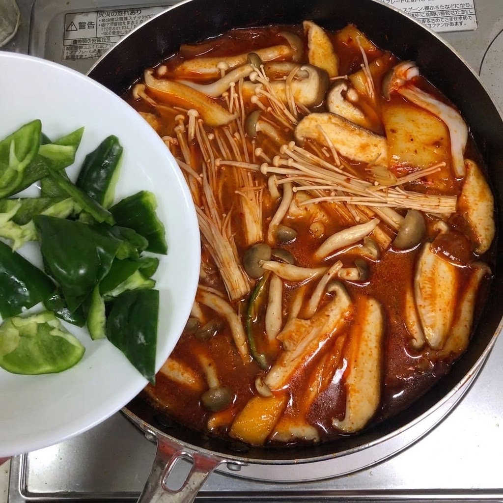 Fette di peperoni verdi aggiunte da un piatto bianco in una pentola di zuppa rossa di kimchi.