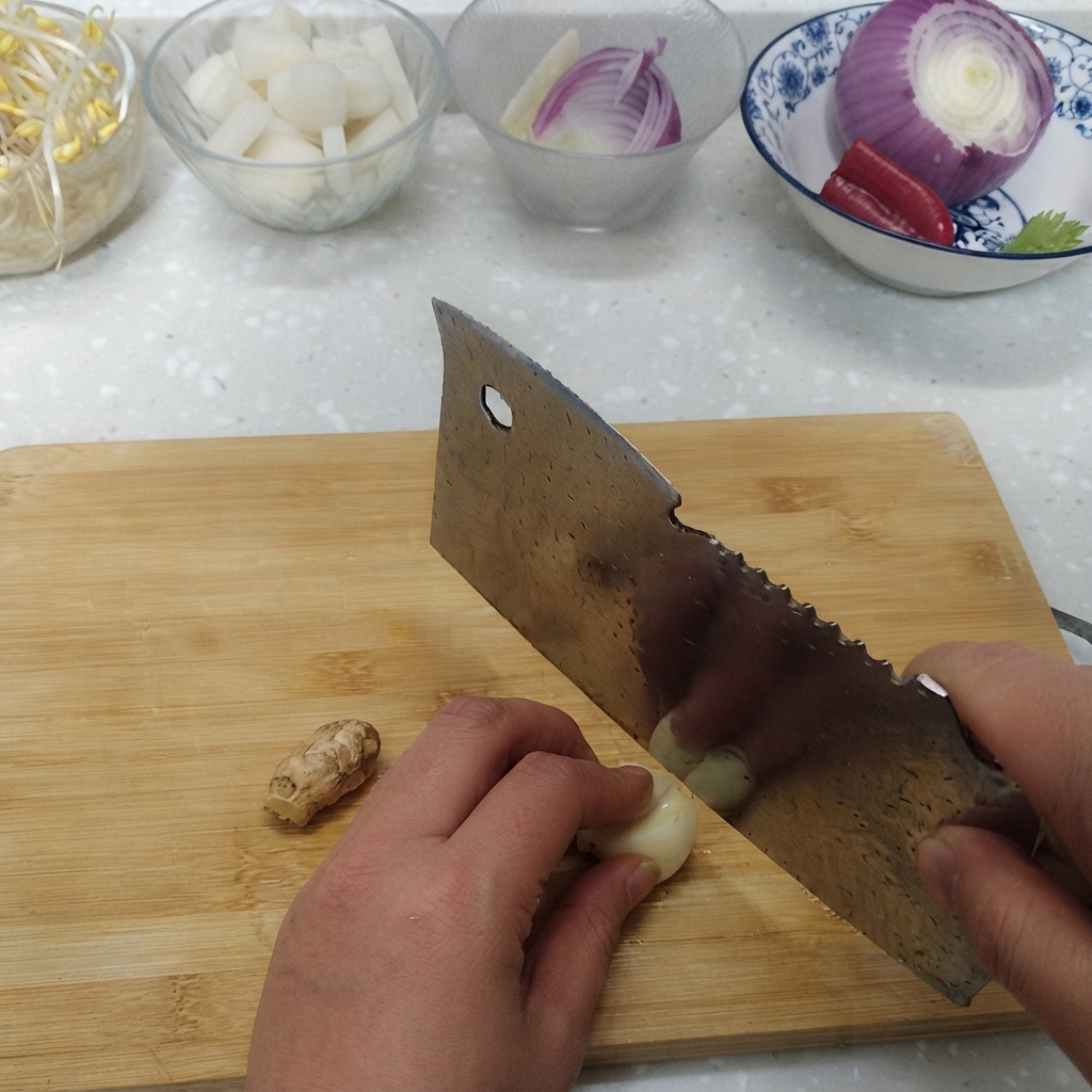 Spicchi daglio e radice di zenzero fresco affettati su un tagliere accanto a ciotole di ingredienti.