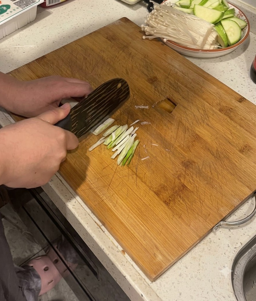 Mani che usano un coltello da cucina per affettare steli di cipollotto su un tagliere di legno.